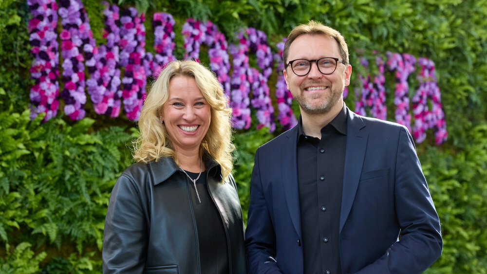 Two people smile in front of a wall of flowers. The woman has curly hair and the man is wearing glasses.