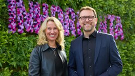 Zwei Menschen lächeln vor einer Blumenwand. Die Frau hat lockiges Haar und der Mann trägt eine Brille.