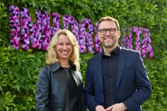 Two people smile in front of a wall of flowers. The woman has curly hair and the man is wearing glasses.