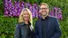 Two people smile in front of a wall of flowers. The woman has curly hair and the man is wearing glasses.