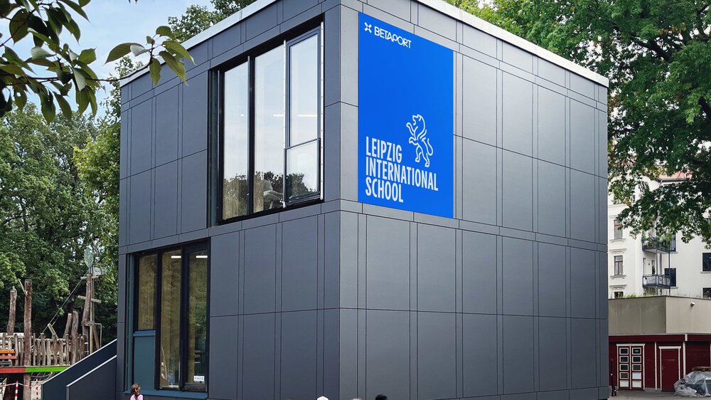 3D-Grafik eines grauen Gebäudes in Würfelform, an dessen Fassade ein blaues Schild mit der Aufschrift „LEIPZIG INTERNATIONAL SCHOOL“ hängt.