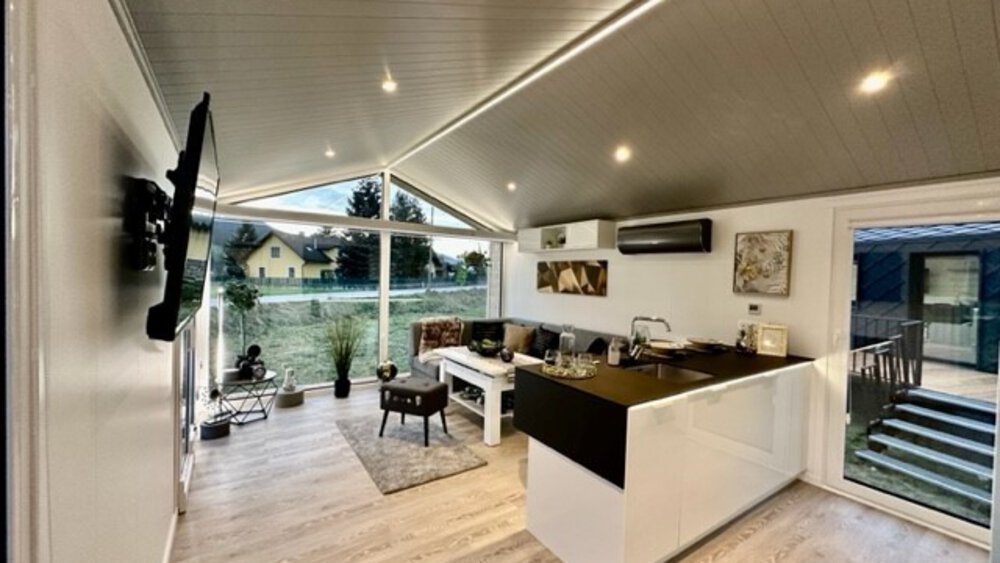 Moderner, offener Wohnbereich mit Küche, Sofa, Wandfernseher und großen Fenstern mit Blick auf den Garten.