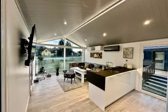 Moderner, offener Wohnbereich mit Küche, Sofa, Wandfernseher und großen Fenstern mit Blick auf den Garten.