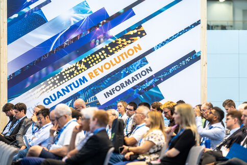 Audience on rows of chairs in an exhibition hall, in front of a large key visual with the inscription 