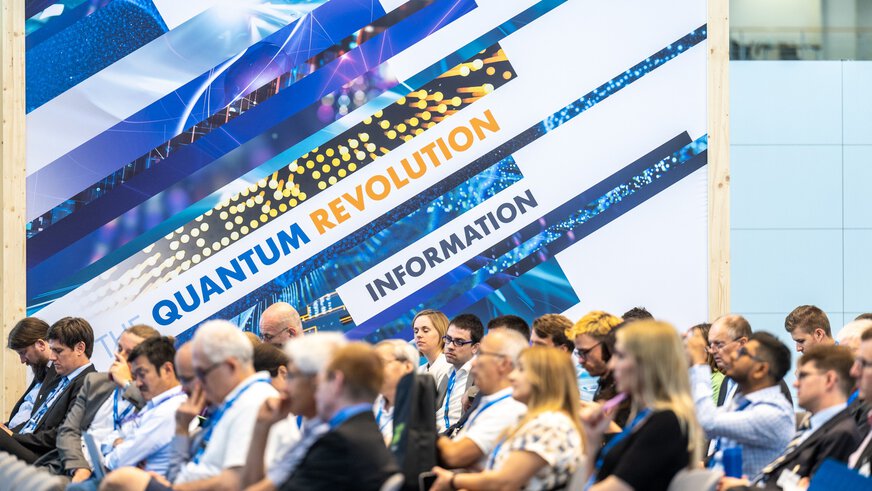 Audience on rows of chairs in an exhibition hall, in front of a large key visual with the inscription 