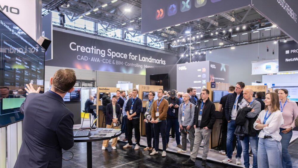 An einem Messestand präsentiert eine Person digitale Lösungen anhand eine Präsentation auf einem großen Bildschirm. Messebesuchende stehen im Halbkreis um den Vortragenden herum.