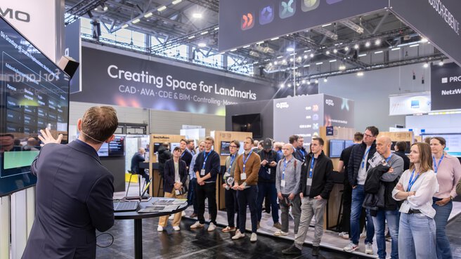 An einem Messestand präsentiert eine Person digitale Lösungen anhand eine Präsentation auf einem großen Bildschirm. Messebesuchende stehen im Halbkreis um den Vortragenden herum.