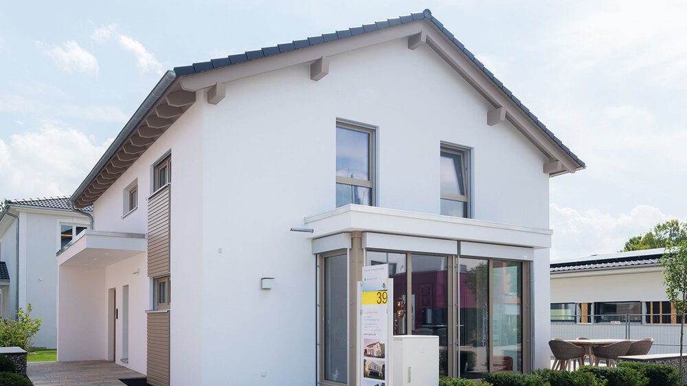 Ein modernes, weißes zweistöckiges Haus mit Satteldach und großen Fenstern, umgeben von Grün.