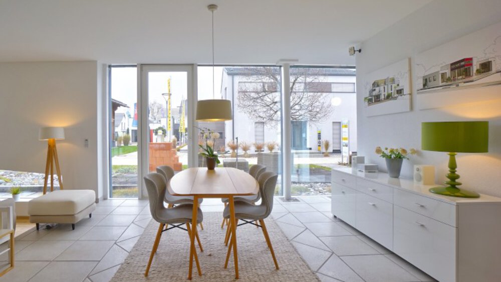 Modernes Esszimmer mit einem Holztisch, grauen Stühlen, einer grünen Lampe und großen Fenstern mit Blick auf einen Garten.