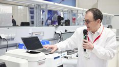 ein Mann in Laborkittel steht vor einem Laptop und liest mit einem Mikrofon Daten aus dem Laptop vor
