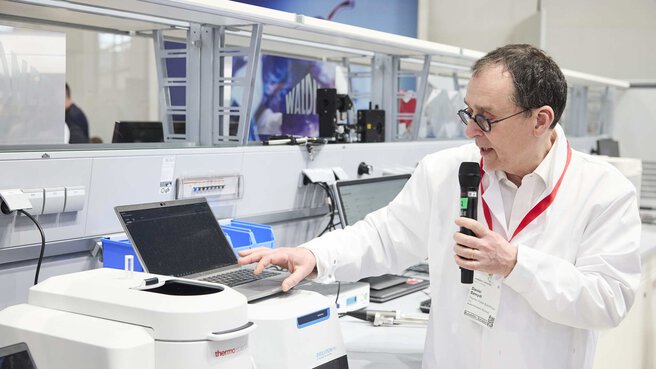 a man in a lab coat stands in front of a laptop and reads out data from the laptop using a microphone