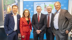 Four men and a woman in suits stand at a transport logistic trade fair stand and pose smiling for a group photo.