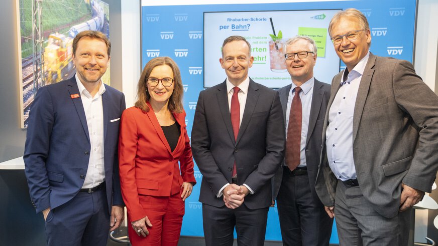 Four men and a woman in suits stand at a transport logistic trade fair stand and pose smiling for a group photo.