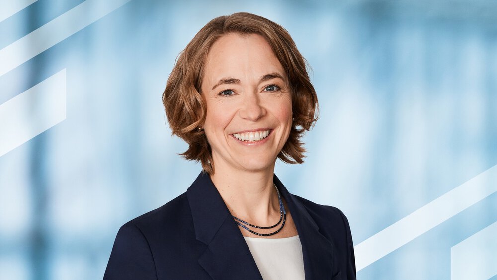 Brightly smiling woman with short, wavy hair in a dark blue suit over a white top.