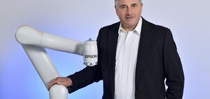  Man in a suit standing next to a collaborative robot arm in a studio environment with a neutral background.