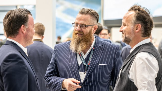 Three men in business attire are smiling and chatting. Other people can be seen behind them.