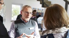 Ein älterer Mann und eine Frau unterhalten sich im Innenbereich. Im Hintergrund sind andere  Menschen und ausgestellte Fotos zu sehen.