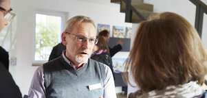 Ein älterer Mann und eine Frau unterhalten sich im Innenbereich. Im Hintergrund sind andere  Menschen und ausgestellte Fotos zu sehen.