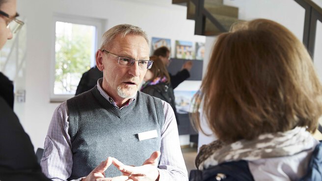 Ein älterer Mann und eine Frau unterhalten sich im Innenbereich. Im Hintergrund sind andere  Menschen und ausgestellte Fotos zu sehen.