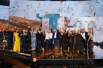 A group of people on stage celebrating amid confetti and holding awards in their hands, against a festive backdrop.