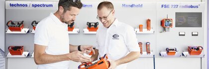 Two men look at an orange radio remote control; in the background, other models are displayed on an exhibitor wall.