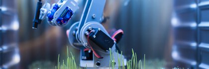 Ein Robotikarm mit einer blauen und schwarzen Struktur ist über mehreren kleinen, transparenten Petrischalen positioniert. In den Schalen wachsen junge Pflanzen, die gerade erst keimen.