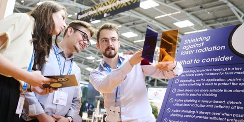 Drei Messebesucher betrachten auf einer Industrie- und Technologiemesse transparente Schutzscheiben und informieren sich an einem Stand über Abschirmung gegen Laserstrahlung.