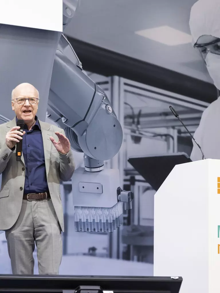 Ein Mann spricht mit Mikrofon auf der Bühne des „MedtecSUMMIT vor einer Wandgrafik mit Roboter und Laborpersonal.