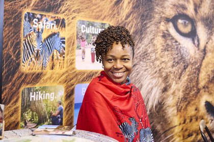 Smiling woman in a red scarf with a safari motif at the Tanzania stand at the f.re.e trade fair, in front of a picture of a lion and information boards about travel.