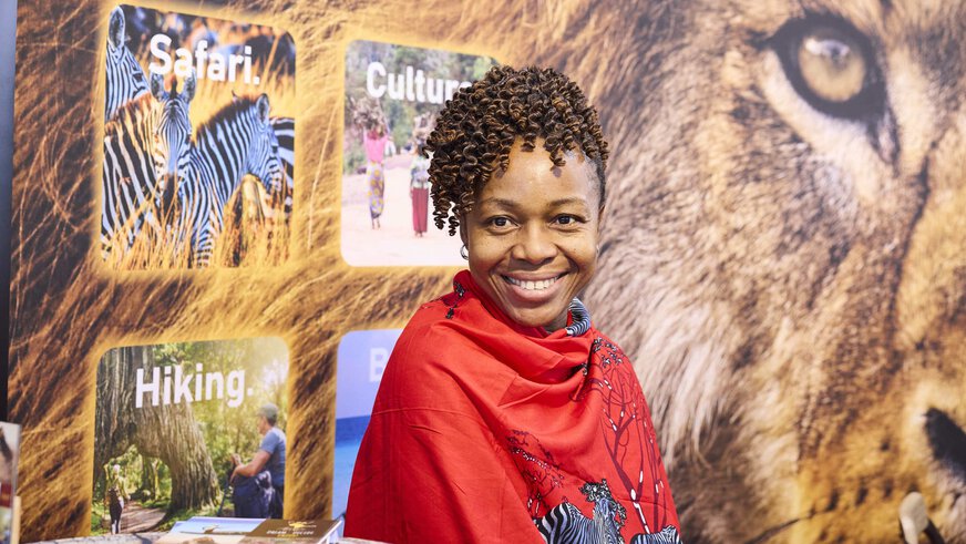 Smiling woman in a red scarf with a safari motif at the Tanzania stand at the f.re.e trade fair, in front of a picture of a lion and information boards about travel.