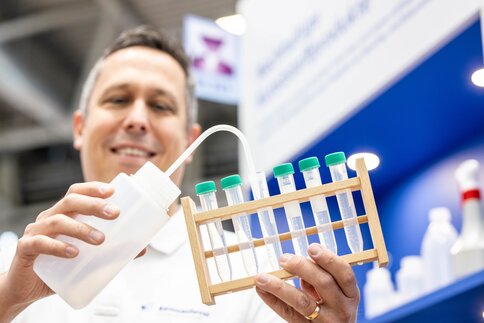 Person holding a wooden rack with several test or centrifuge tubes with green caps, filling one tube with liquid from a squeeze bottle.