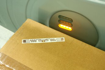 Ein Paket mit einem Aufkleben neben einem RFID Scanner, der ein orangefarbenes Licht anzeigt.
