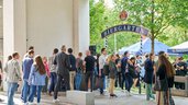 People stream through the entrance to a beer garden, with trees and blue parasols in the background.