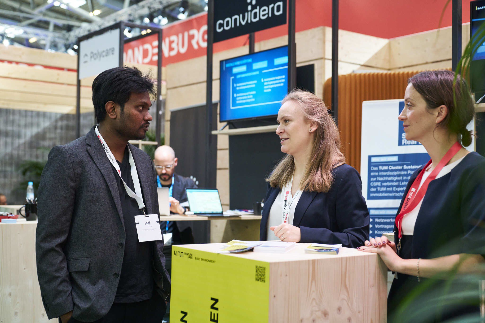 Three people in business attire stand around a small wooden counter at an exhibition stand and talk.