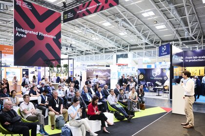 Lecture at the Laser World of Photonics trade show in front of an audience, with exhibition booths and a sign reading “Integrated Photonics Area” in the background
