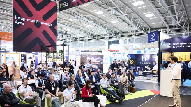 Lecture at the Laser World of Photonics trade show in front of an audience, with exhibition booths and a sign reading “Integrated Photonics Area” in the background