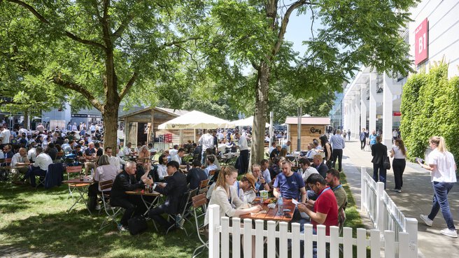 Biergarten im Atrium der Messe München mit vielen Besuchern, die an Tischen sitzen und essen.