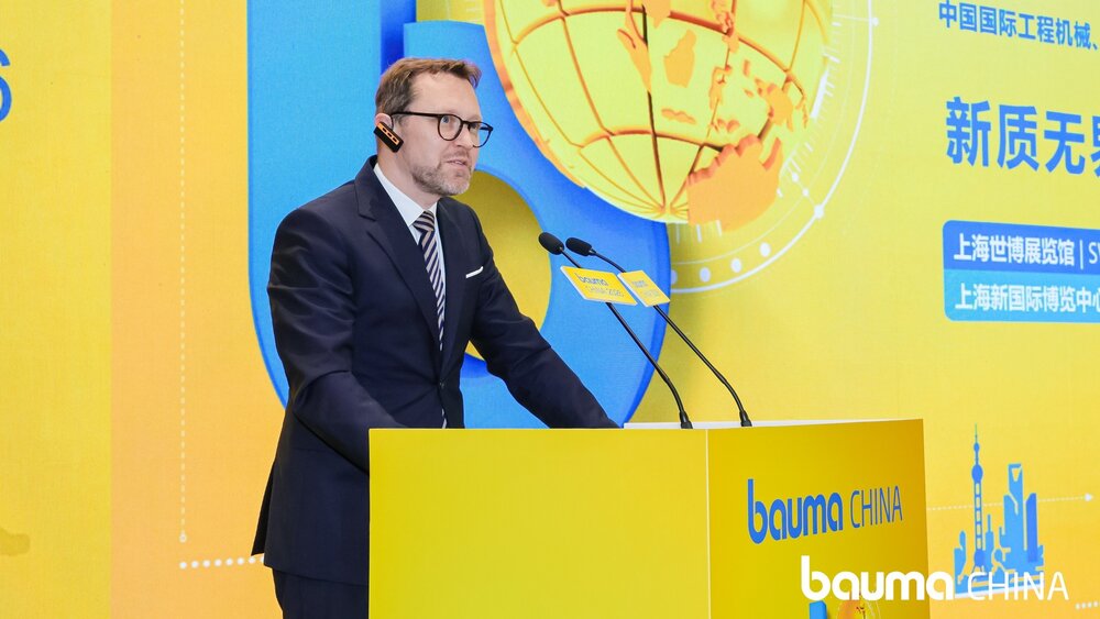 A man in a suit and glasses is giving a speech at a yellow lectern bearing the ‘bauma CHINA’ logo, against a bright yellow and blue background and a large image of a globe.