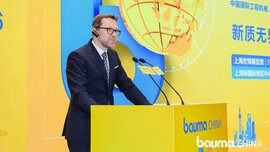 A man in a suit and glasses is giving a speech at a yellow lectern bearing the ‘bauma CHINA’ logo, against a bright yellow and blue background and a large image of a globe.