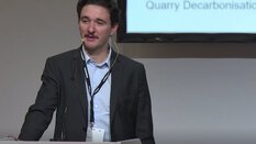 Ein Sprecher mit Headset-Mikrofon am Podium, im Hintergrund ist Text auf einem Bildschirm zu sehen.