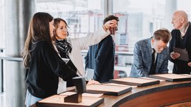 Five people are standing around a semicircular counter, looking at various pieces of jewelry displayed in black boxes.