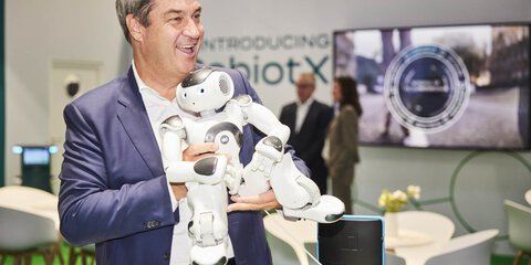 Lachender Mann im Anzug, der an einem Messestand einen humanoiden Roboter auf dem Arm trägt.