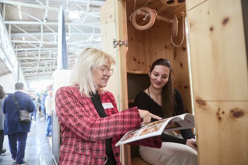 A young woman sits in a wooden booth at an exhibition stand and an older woman explains something to her while pointing to a catalog