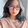 A person with long, wavy black-and-white hair, wearing glasses and a peach-colored top, looks into the camera in natural light.