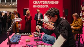 Ein Mann arbeitet an einem rosafarbenen Tisch während einer Handwerksveranstaltung an Schmuck; im Hintergrund schauen Leute zu.