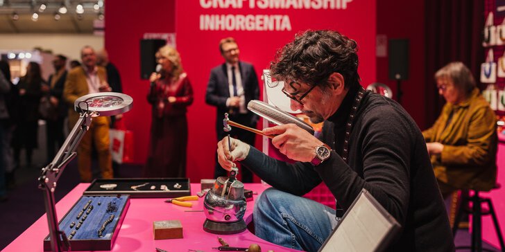 Ein Mann arbeitet an einem rosafarbenen Tisch während einer Handwerksveranstaltung an Schmuck; im Hintergrund schauen Leute zu.