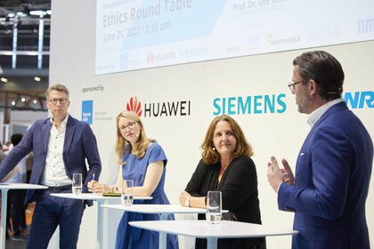 Vier Personen beteiligen sich an einer Podiumsdiskussion auf einer Bühne, vor einer Logowand mit einer Projektionsfläche.