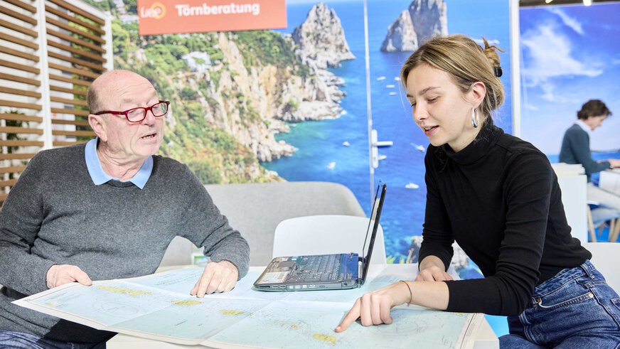 A man and a woman discuss a nautical chart during a cruise consultation at the f.re.e trade fair, in front of a maritime background image.