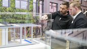 Two men are looking at a display case at an exhibition stand in which a building model with several ceiling and floor examples is presented.