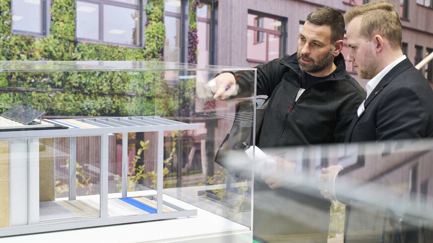Zwei Männer betrachten eine Vitrine an einem Messestand, in der ein Gebäudemodell mit mehreren Decken- und Bodenbeispielen präsentiert wird.